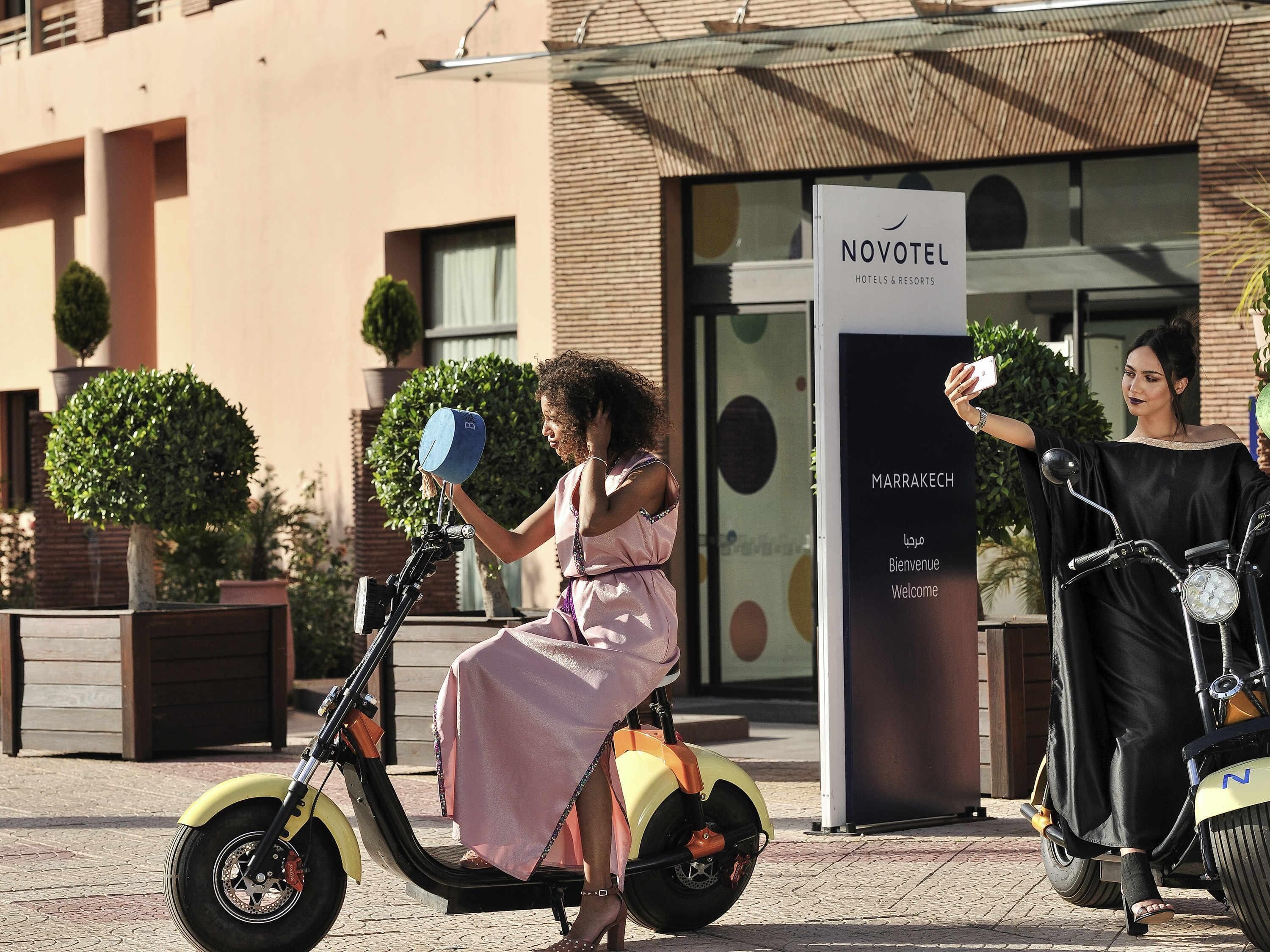 A person on a yellow electric scooter in front of a hotel with a sign that reads 'NOVOTEL HOTELS & RESORTS' and 'MARRAKECH' indicating the location.