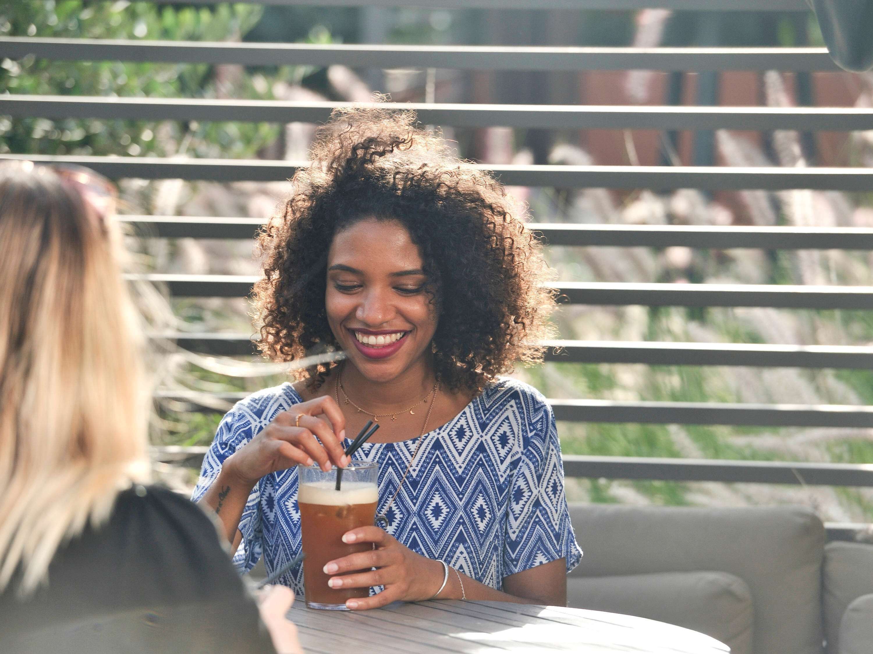 A person sitting at an outdoor table with a drink, surrounded by a railing and greenery.
