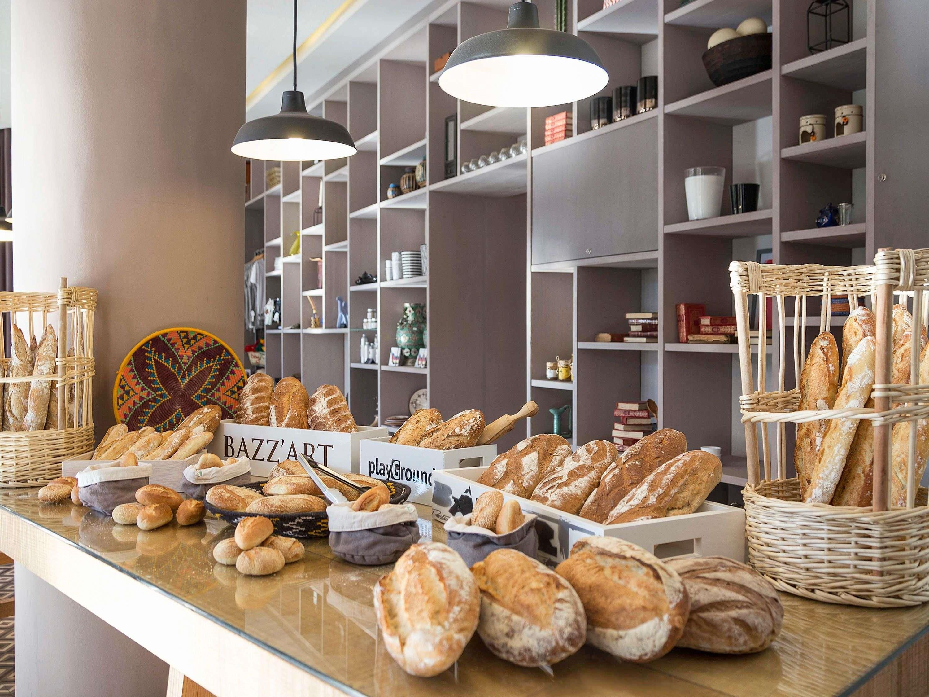 Mostrador de panadería con panes y pasteles varios, estantes con utensilios de cocina e iluminación colgante.