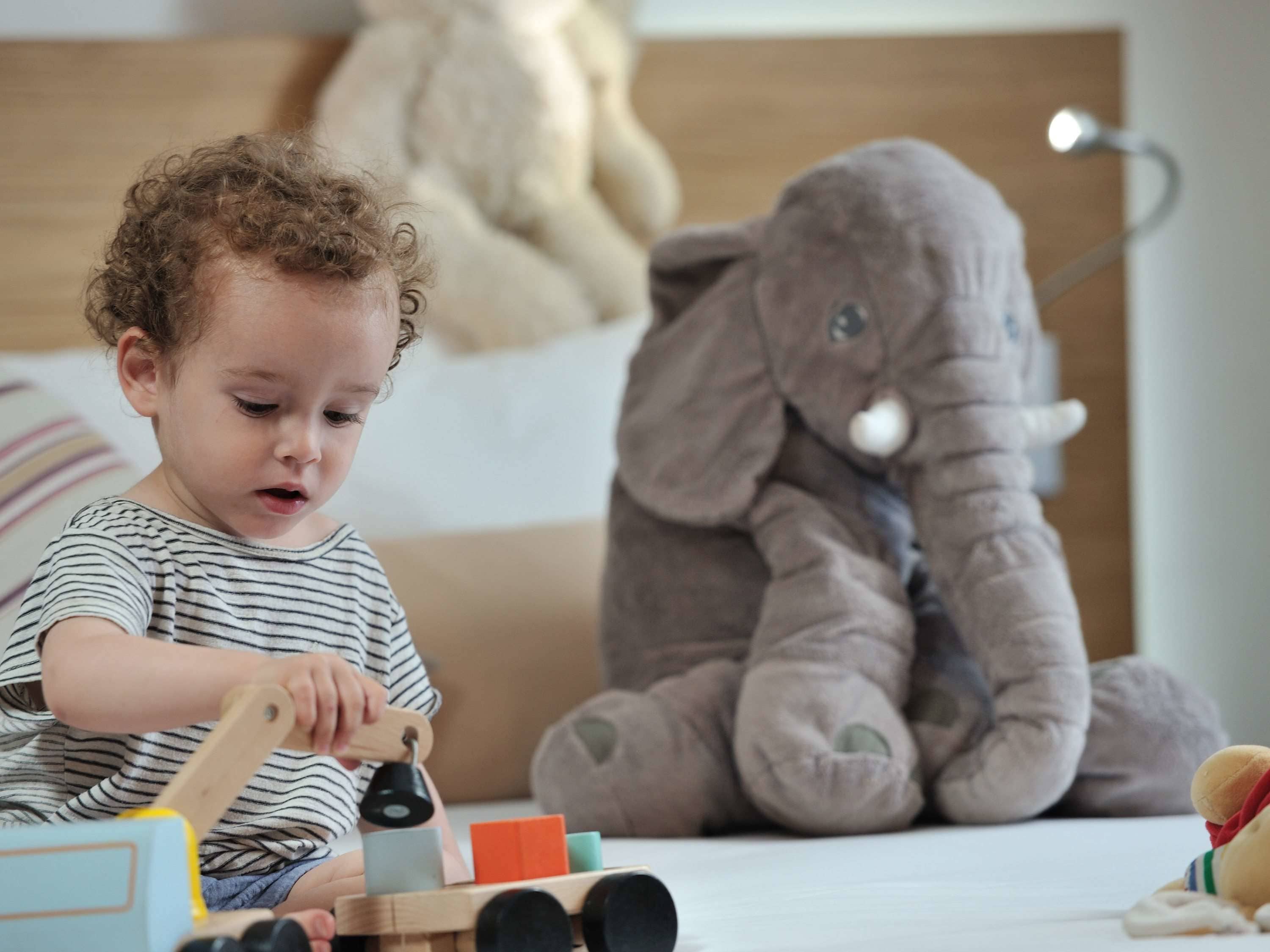 Niño jugando con juguetes en una cama, con un elefante de peluche y una lámpara al fondo.