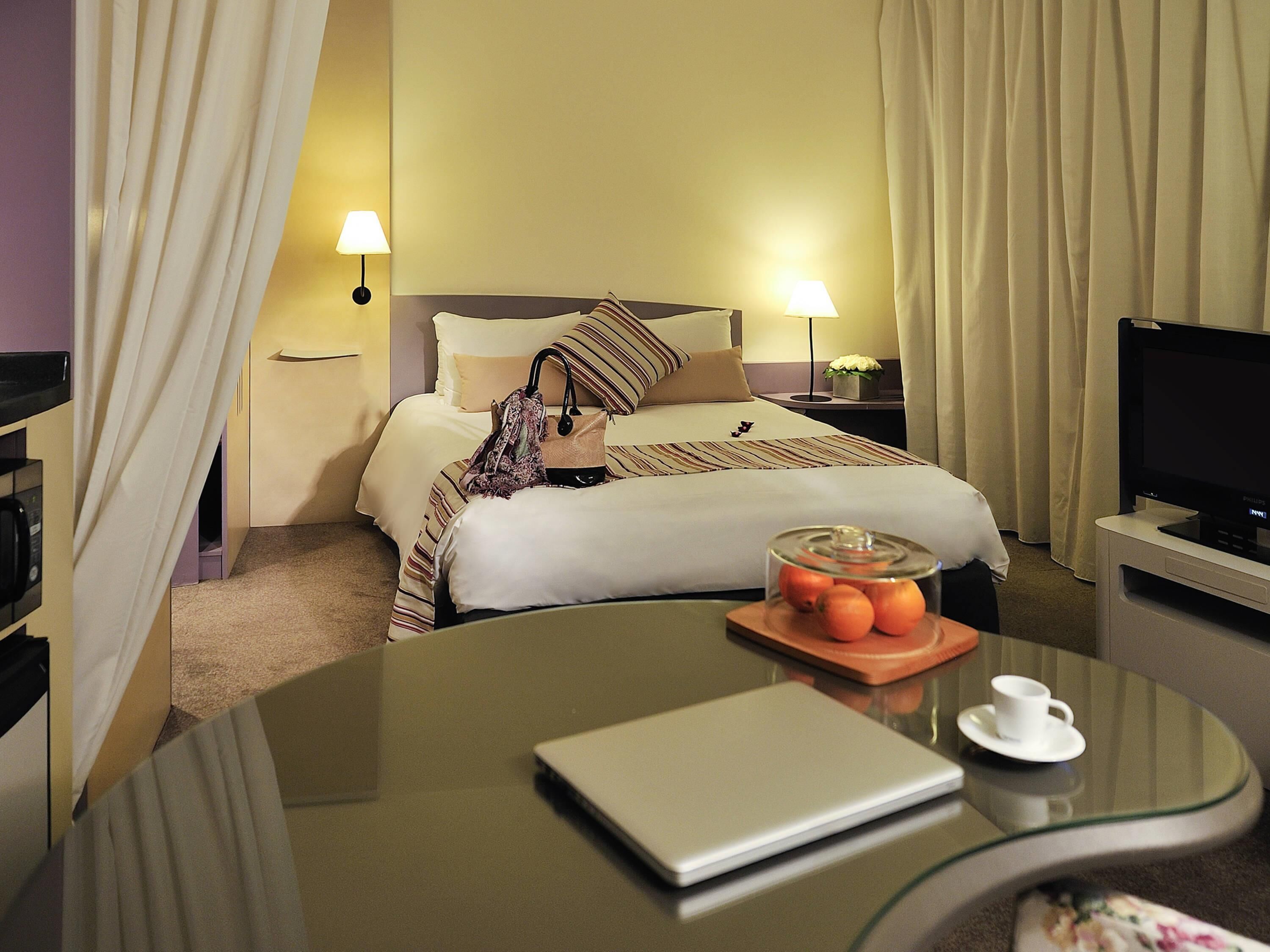 A hotel room with a bed, bedside lamps, a TV, a glass table with a laptop, a cup, and a fruit bowl.