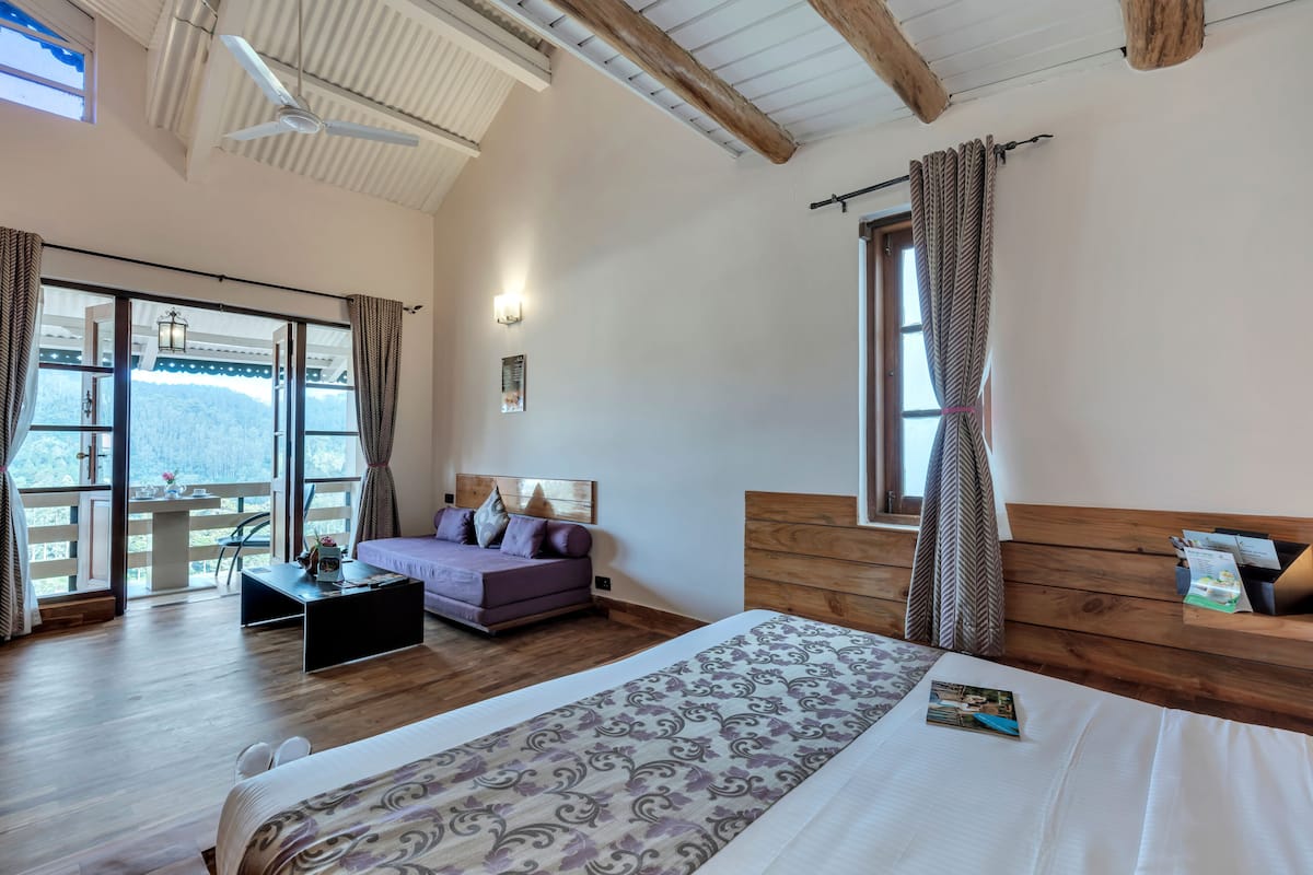 A spacious bedroom with a large bed, a wooden headboard, a sofa, and a balcony with a view of the mountains.