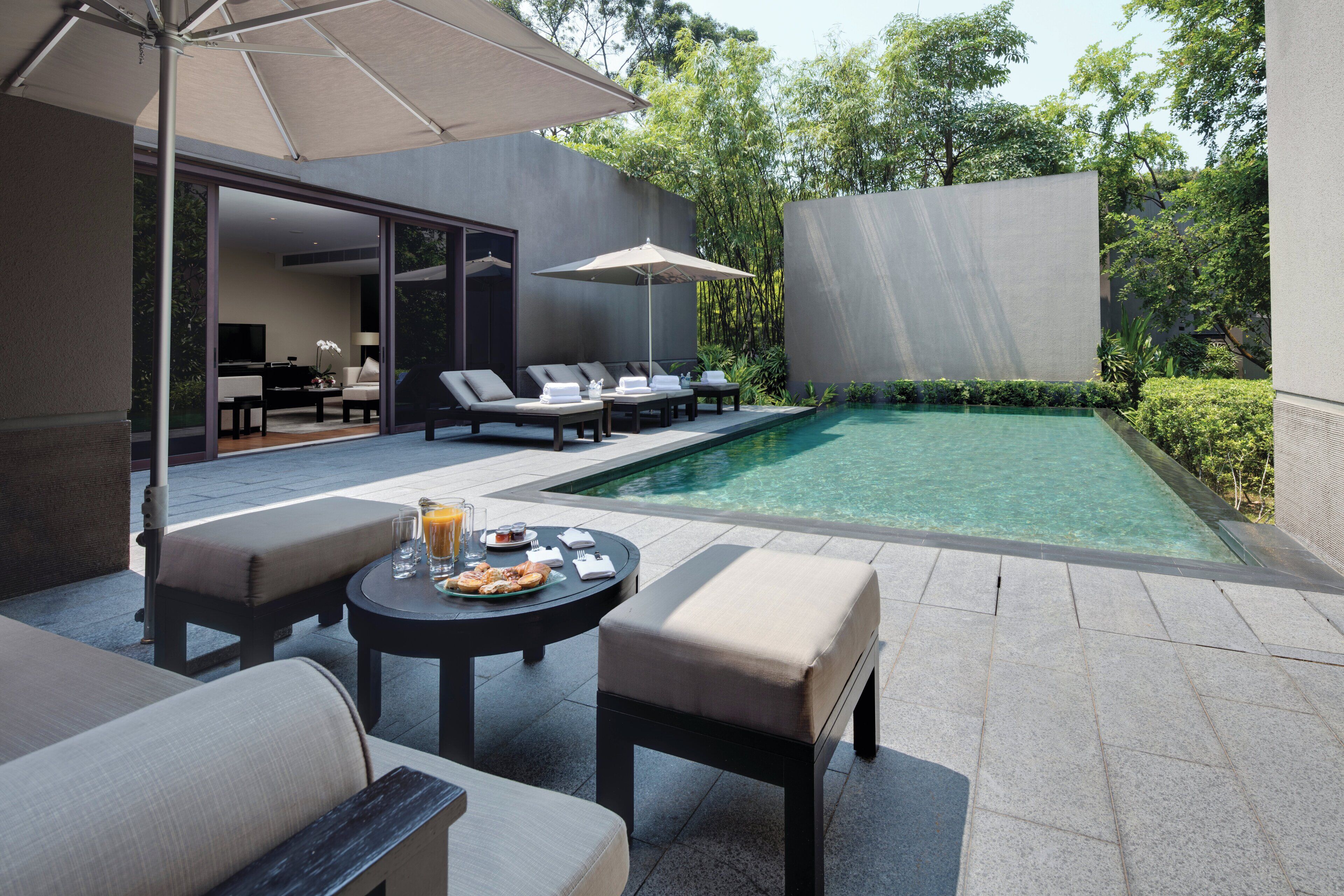 Zona de piscina exterior moderna con sillones reclinables, una mesa con desayuno y un paraguas grande.