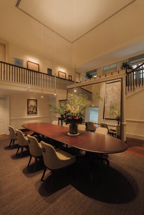 Una amplia sala de conferencias con una gran mesa de madera, varias sillas y un arreglo floral decorativo. Se ve una escalera con barandilla al fondo.