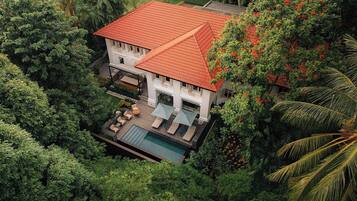 A two-story house with a red tiled roof, surrounded by lush greenery and a swimming pool.