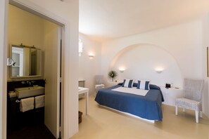 Ein Schlafzimmer mit Bett, Sessel, Nachttisch, Spiegel und einem Badezimmer mit Waschbecken und Handtuchhalter.