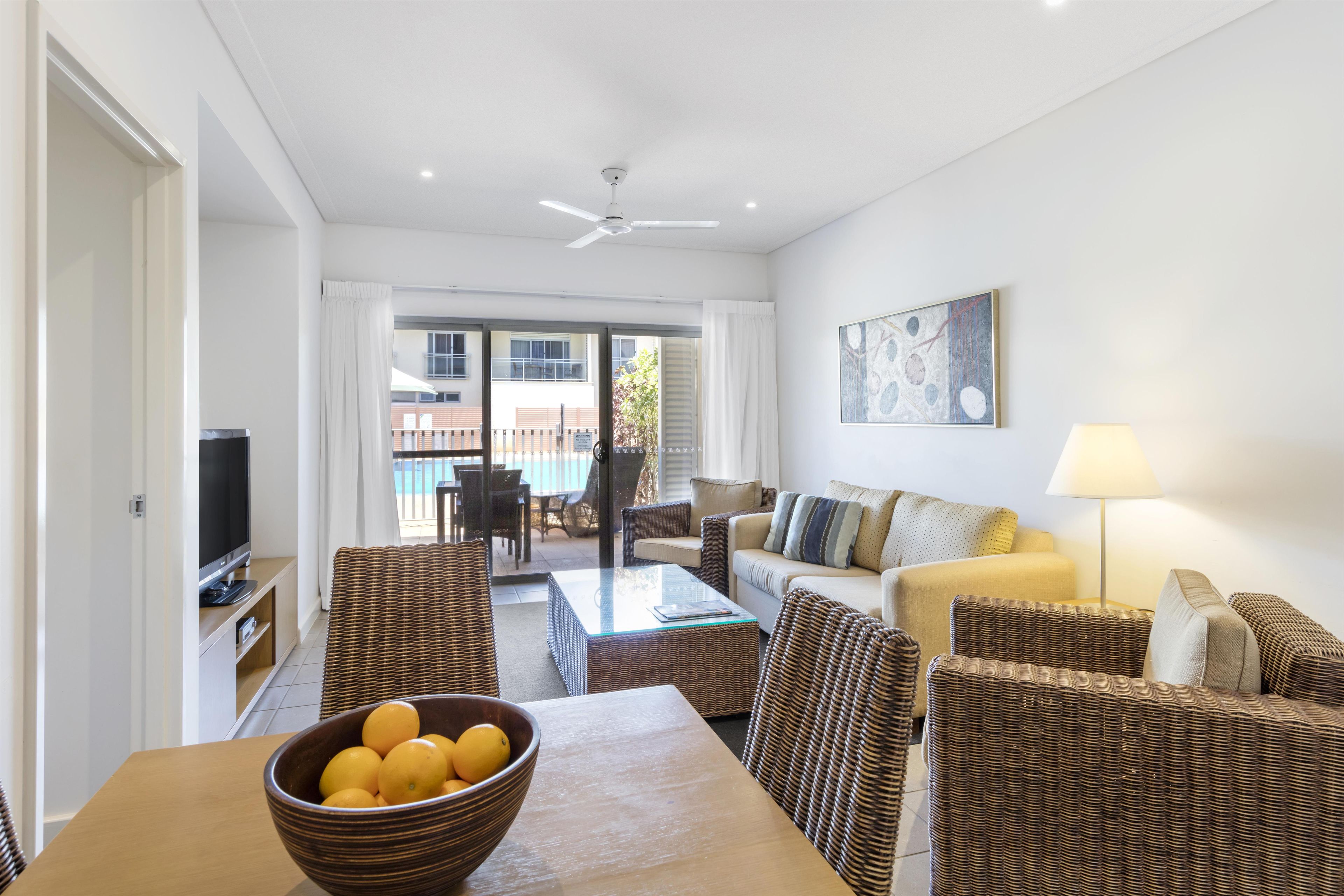 A modern living room with a flat-screen TV, wicker furniture, a glass coffee table, and a view of a pool and buildings outside.