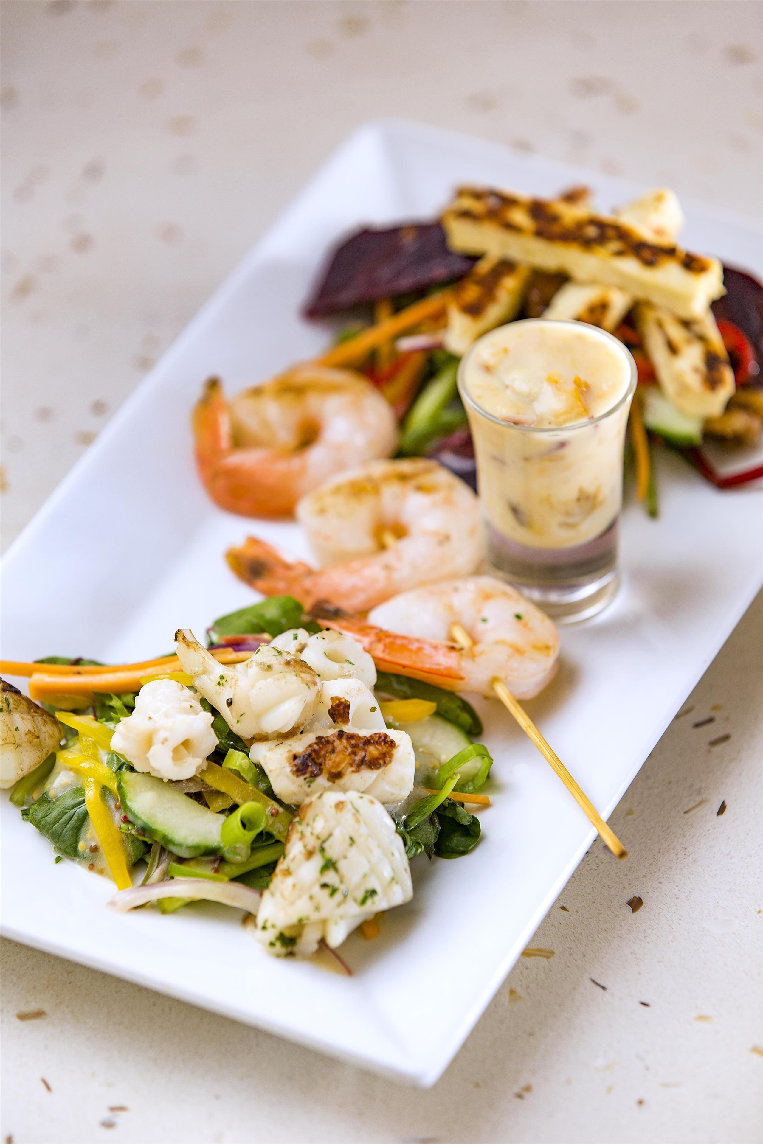 A white rectangular plate with a variety of seafood, vegetables, and a glass of a creamy beverage.
