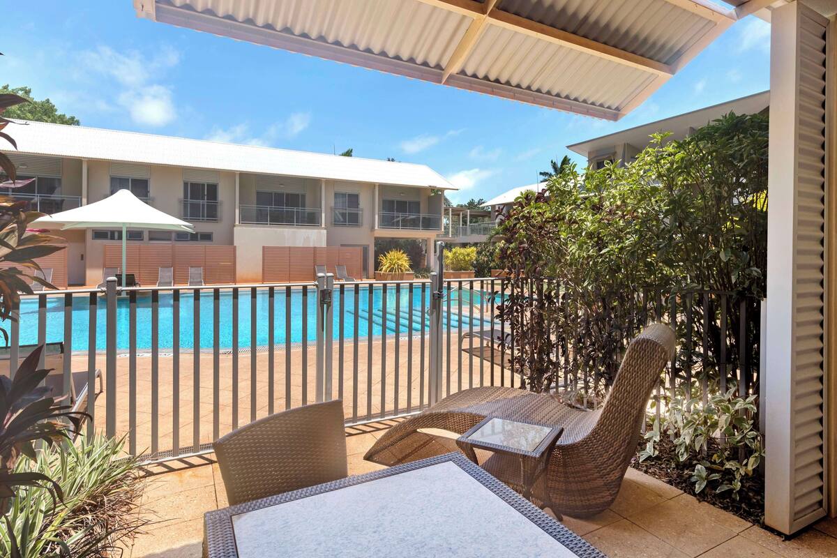 A covered outdoor area with a pool, wicker furniture, and a view of a building in the background.
