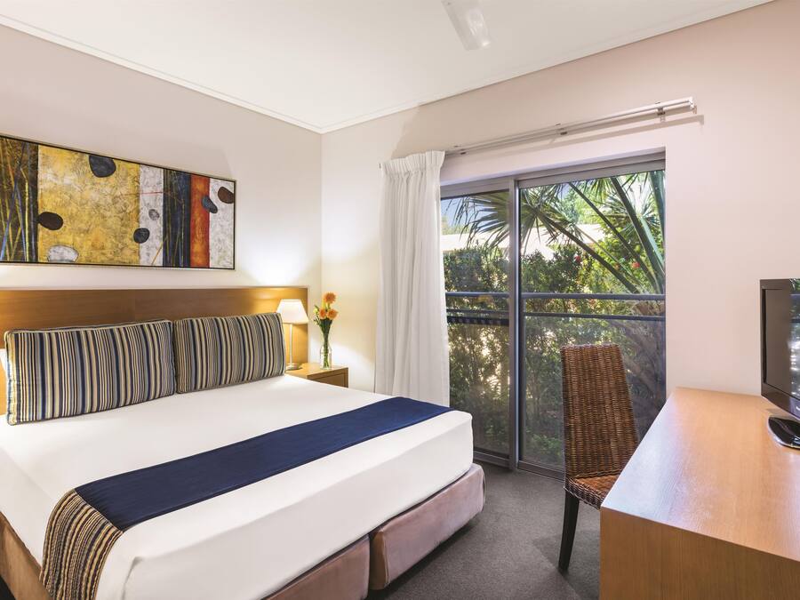 Une chambre d’hôtel avec un lit, un bureau avec une télévision, une chaise, une fenêtre donnant sur des arbres et un tableau au mur.