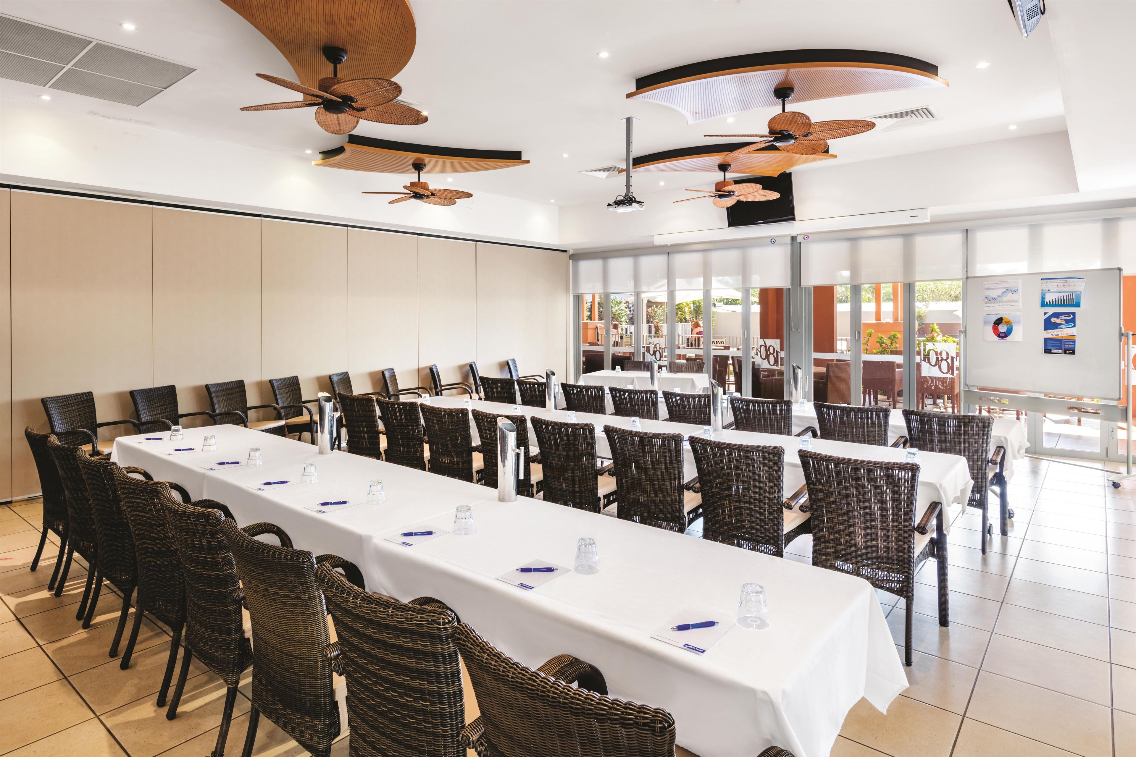 A conference room with long tables, wicker chairs, ceiling fans, and a view of the outdoors.