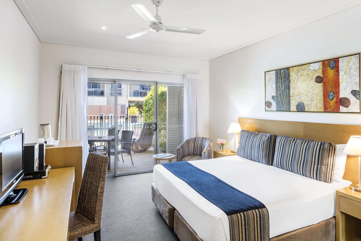 Habitación de hotel con cama, escritorio con televisor, silla, ventilador de techo y vistas al exterior.