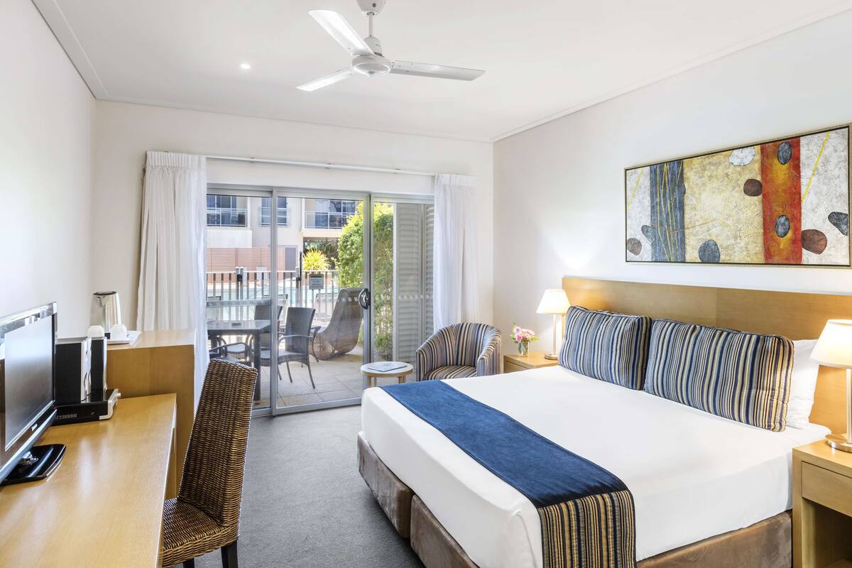 A hotel room with a bed, a desk, a chair, a television, a ceiling fan, and a view of the outside.
