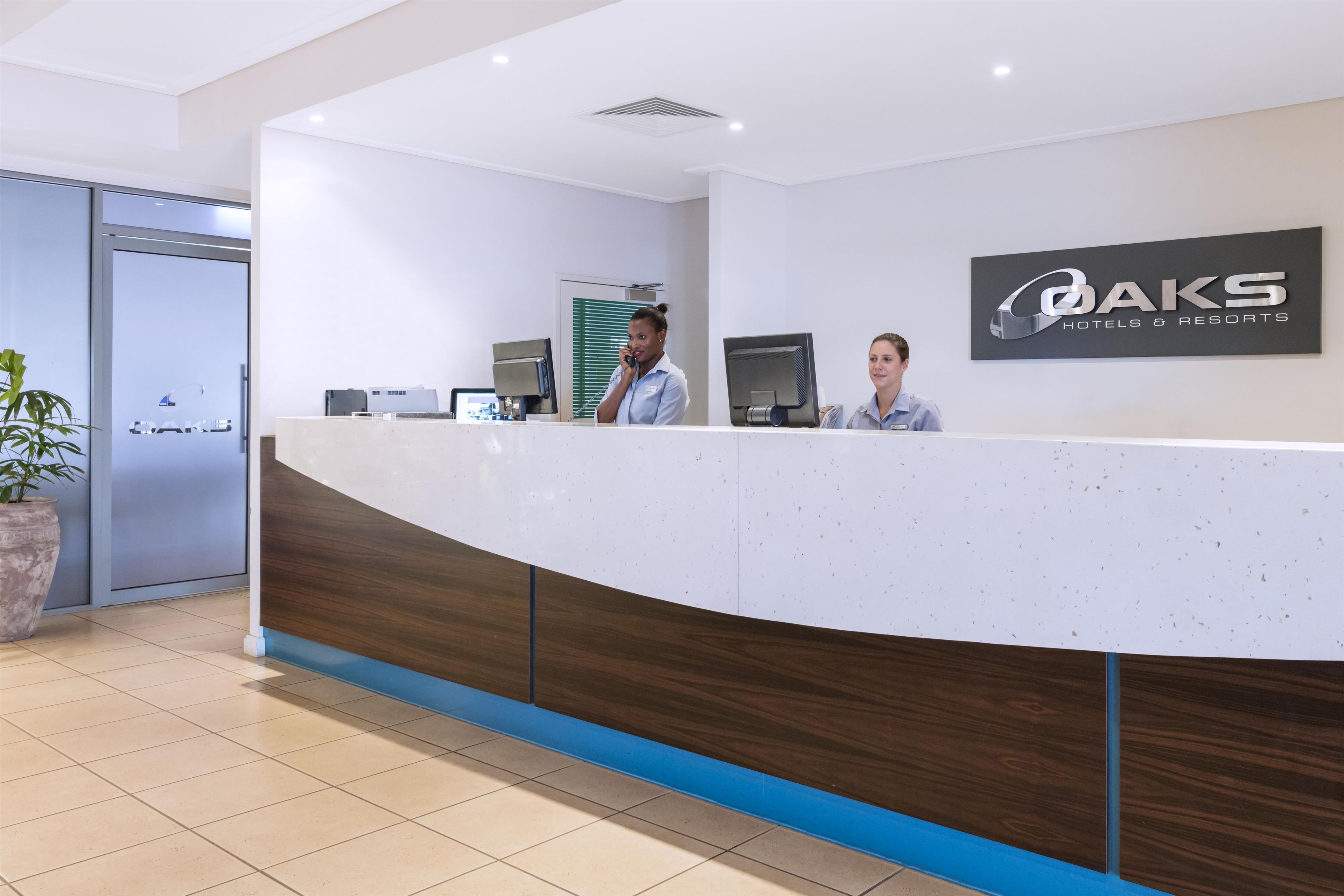 A reception desk with a logo for OAKS Hotels & Resorts, two staff members, a large potted plant, and a door to the left.