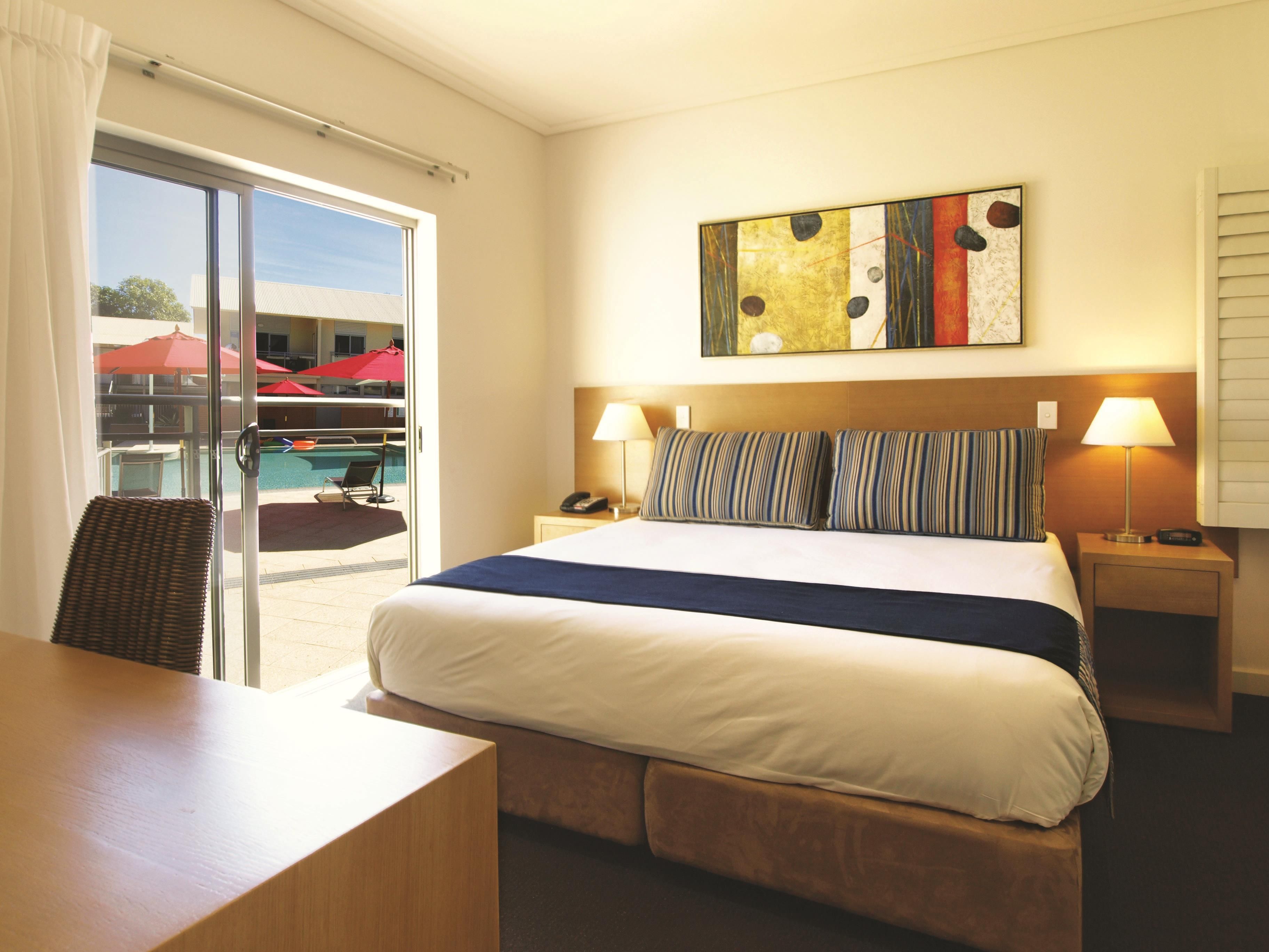 A hotel room with a bed, a desk, a chair, a bedside table with a lamp, a phone, and a painting on the wall.