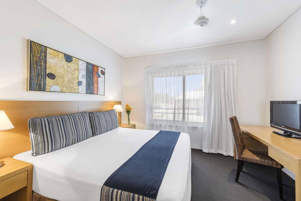 A hotel room with a bed, a desk, a television, a painting, and a window with curtains.