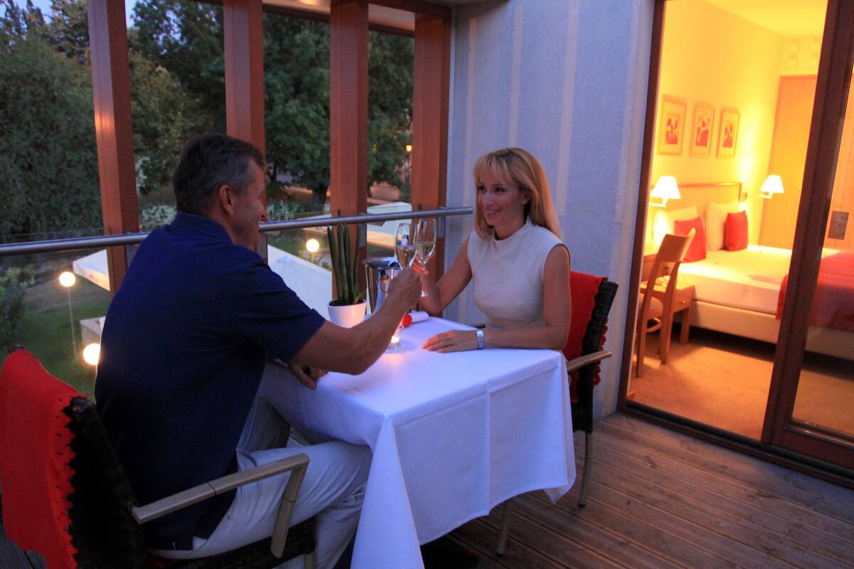 A couple seated at an outdoor table with a white tablecloth, a potted plant, and a view of a bedroom with a bed and a nightstand.