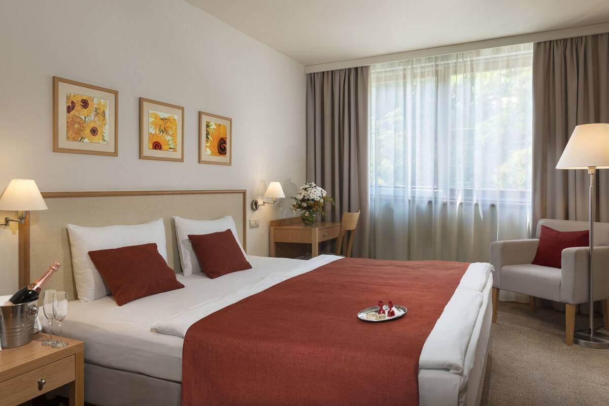 A hotel room with a bed, two bedside tables, a chair, a desk, and a window with curtains.
