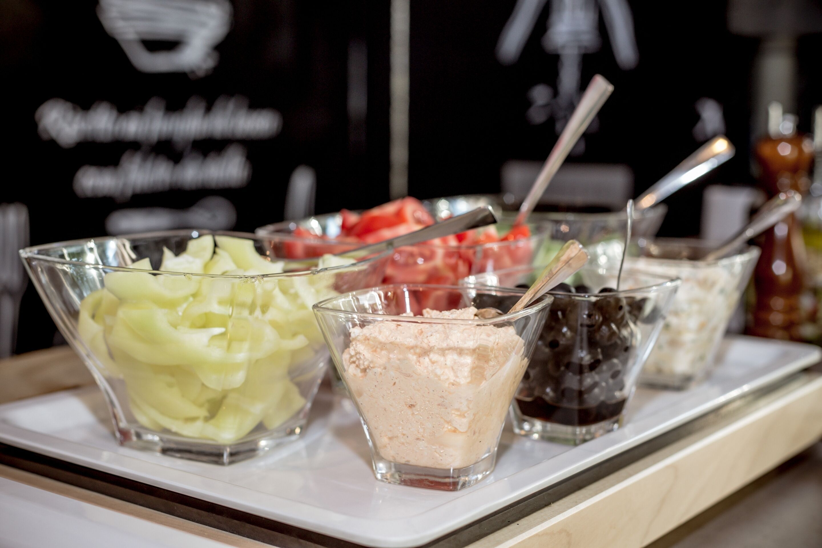 A buffet with various food items including sliced cucumbers, a pink dessert, and a dark dessert in clear glasses.