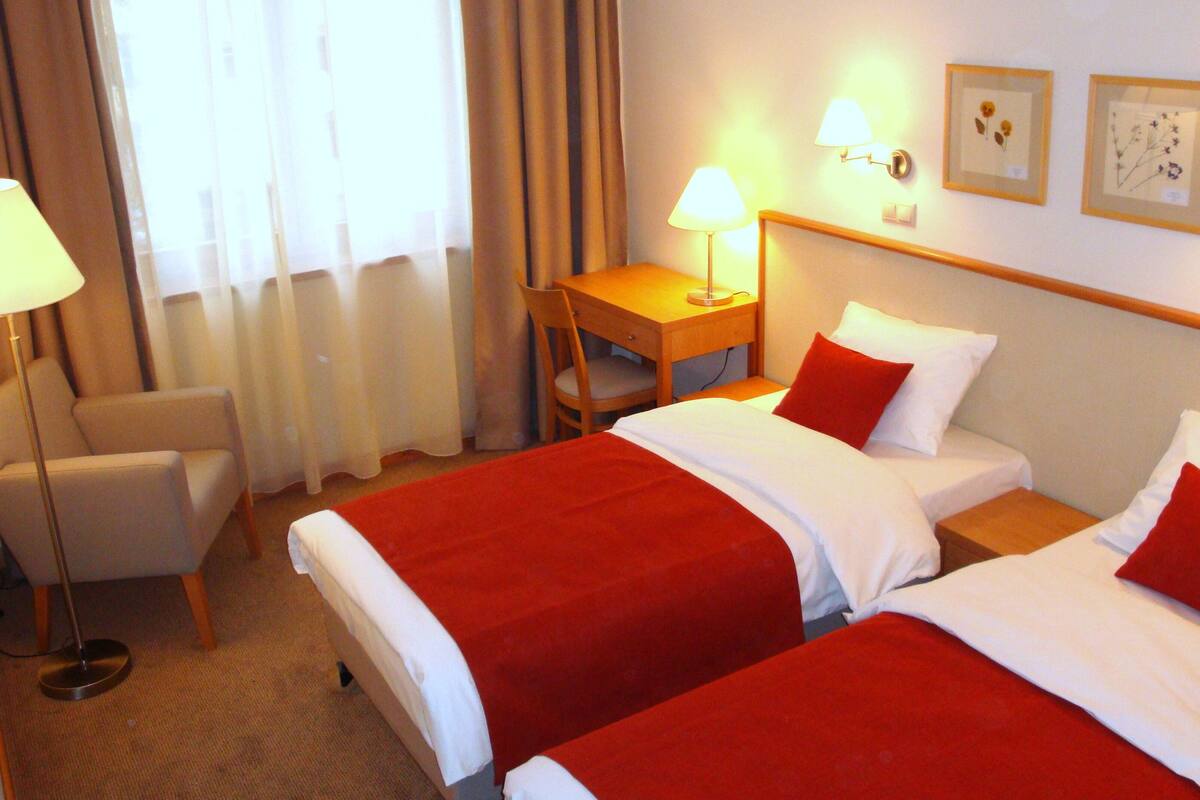 A hotel room with two beds, a desk, a chair, and a lamp.