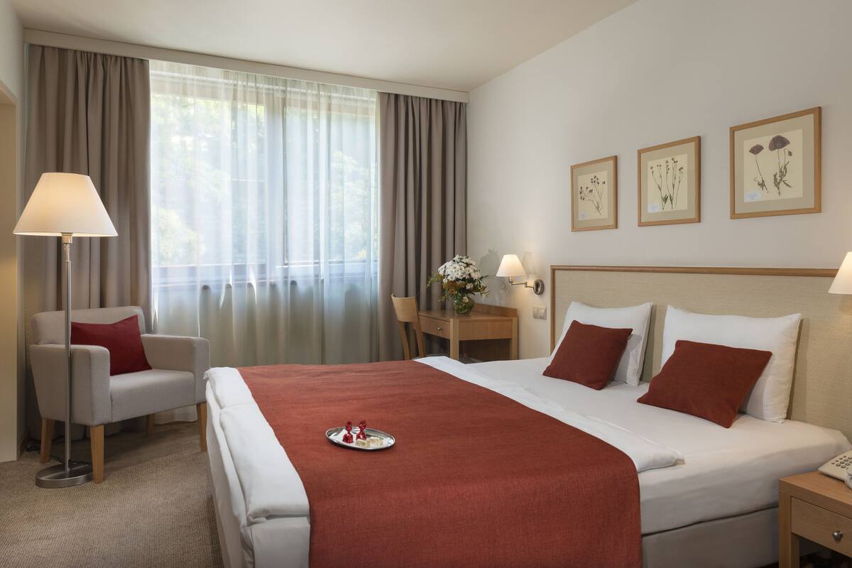 A hotel room with a bed, bedside lamps, a desk, a chair, and a window with curtains.