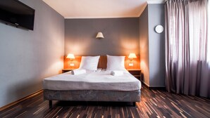 A hotel room with a bed, two bedside lamps, a TV mounted on the wall, and a window with curtains.