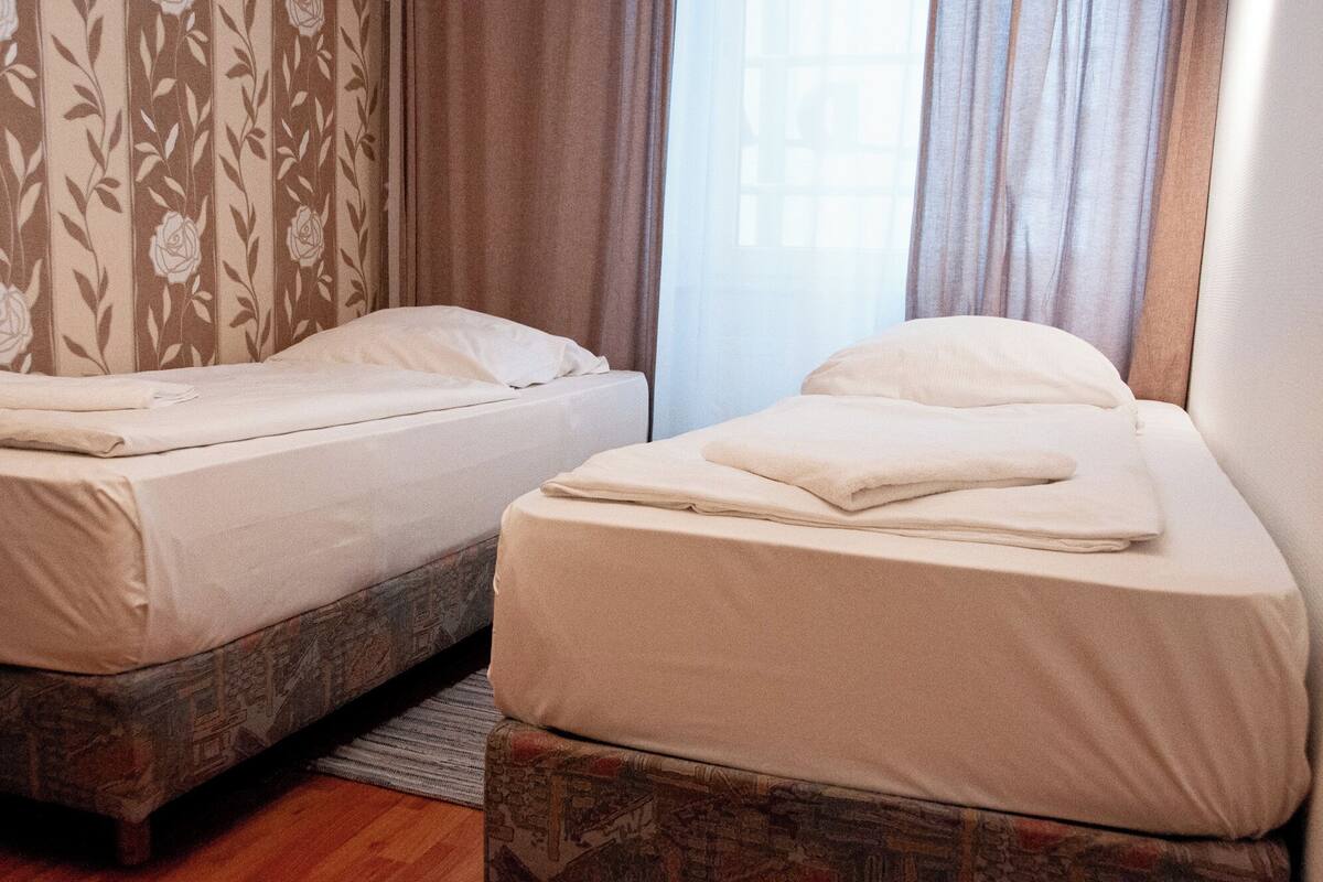 Two single beds with white linens in a room with patterned wallpaper and a window with curtains.