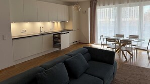 A modern kitchen with white cabinets, a dining area with a glass table and chairs, and a grey sofa.