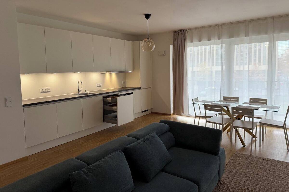 A modern kitchen with white cabinets, a dining area with a glass table and chairs, and a grey sofa.