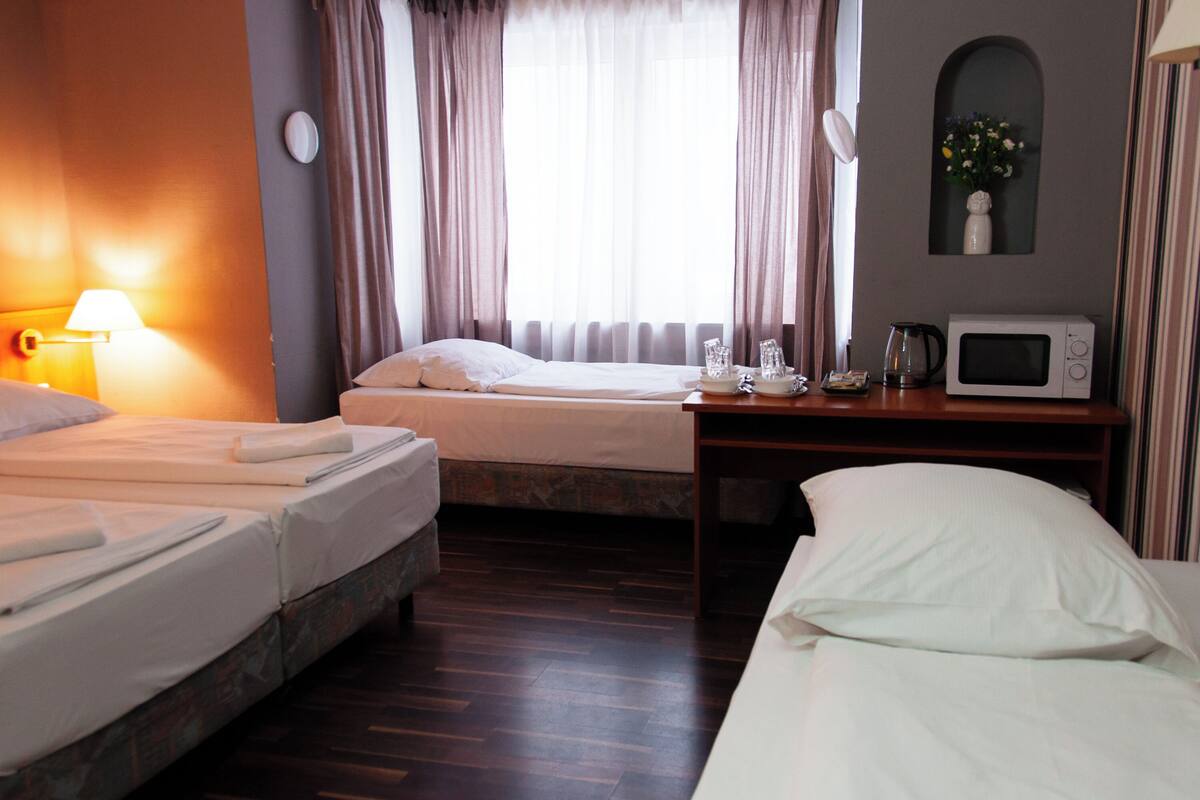 A hotel room with two beds, a bedside table with a microwave, a clock, and a window with curtains.