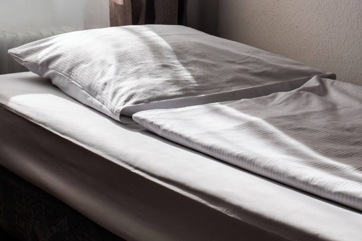 A single bed with a white mattress and pillows, a window with curtains, and a plain wall.
