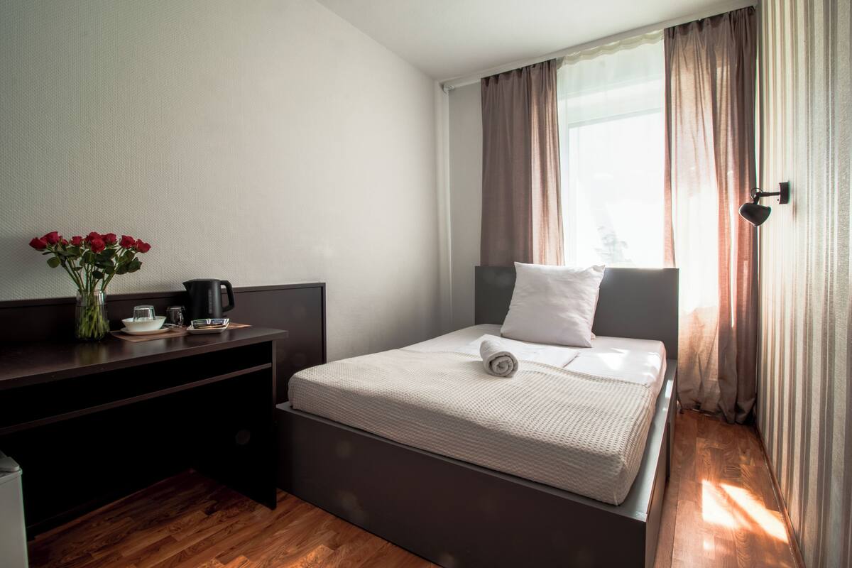 A neatly made bed with a white mattress, a small bedside table with a vase of red flowers, and a coffee maker.