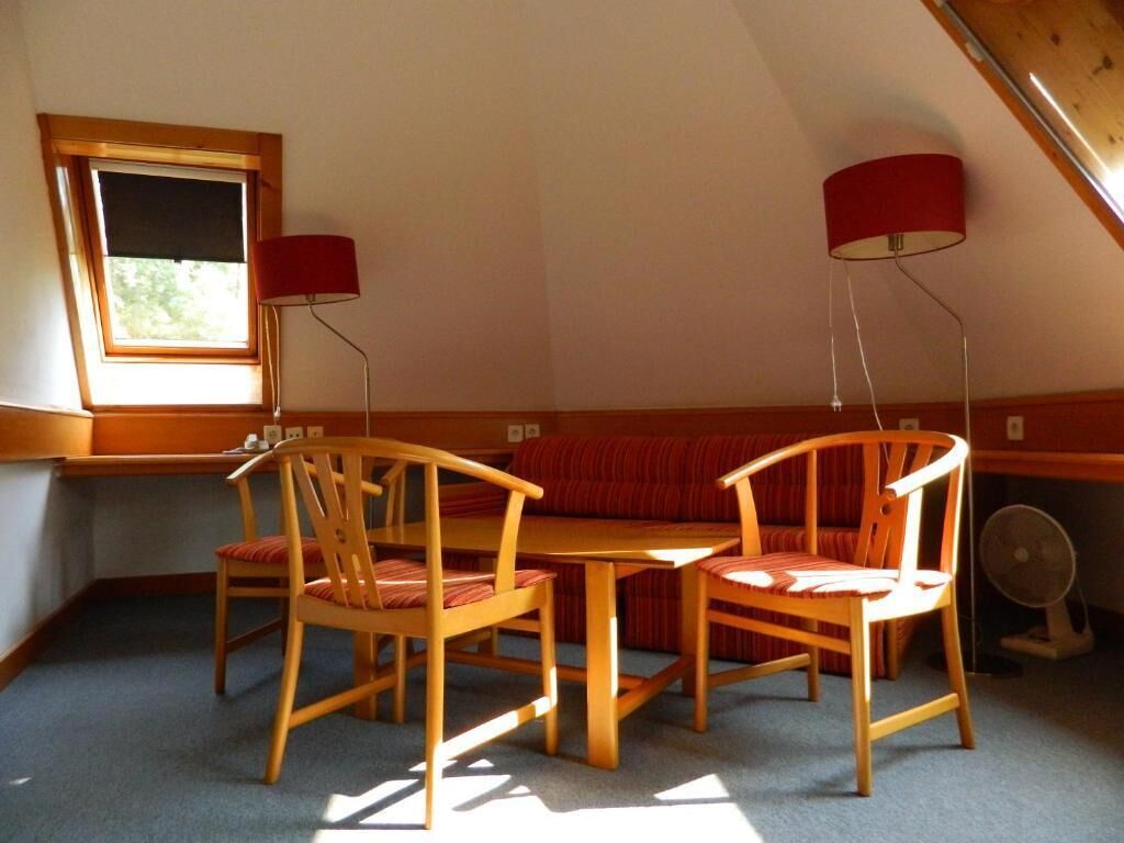 A room with a sloped ceiling, a wooden table, two chairs, a red sofa, and a window with blinds.