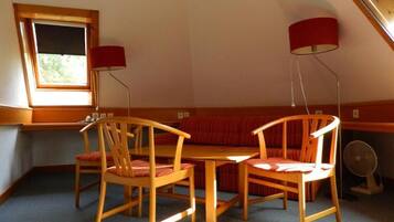 A room with a sloped ceiling, a wooden table, two chairs, a red sofa, and a window with blinds.