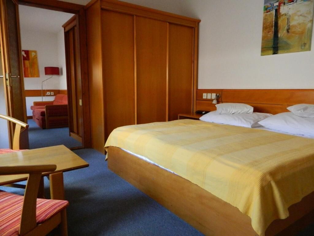 A hotel room with a bed, a wooden wardrobe, a desk with a chair, and a painting on the wall.