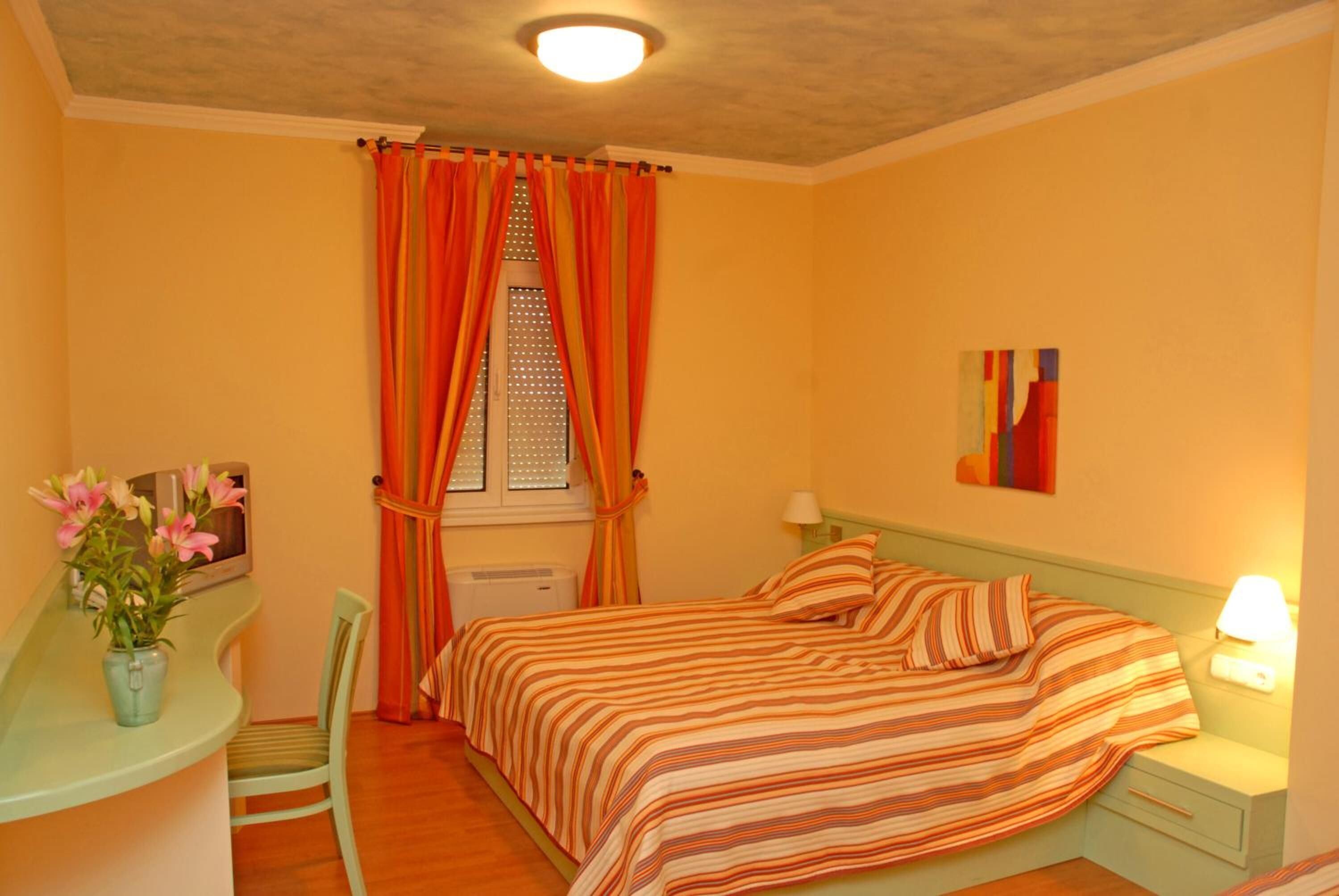 A bedroom with a bed, a desk with a chair, a window with curtains, and a nightstand with a lamp.