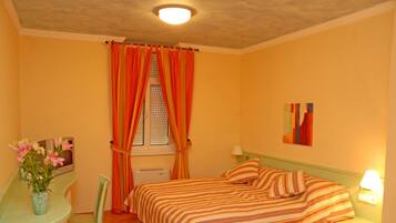 A bedroom with a bed, a desk with a chair, a window with curtains, and a nightstand with a lamp.