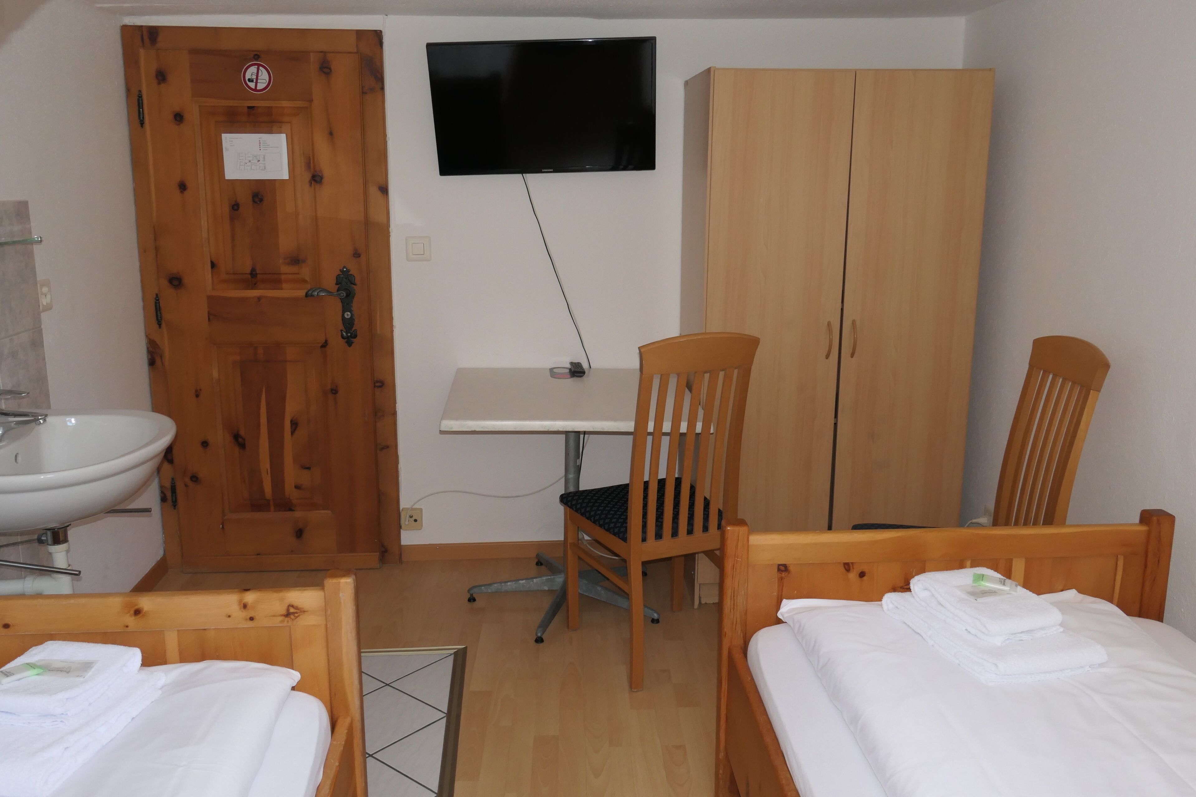 A small, clean room with a wooden door, a TV mounted on the wall, a desk, a chair, a bed, and a wardrobe.