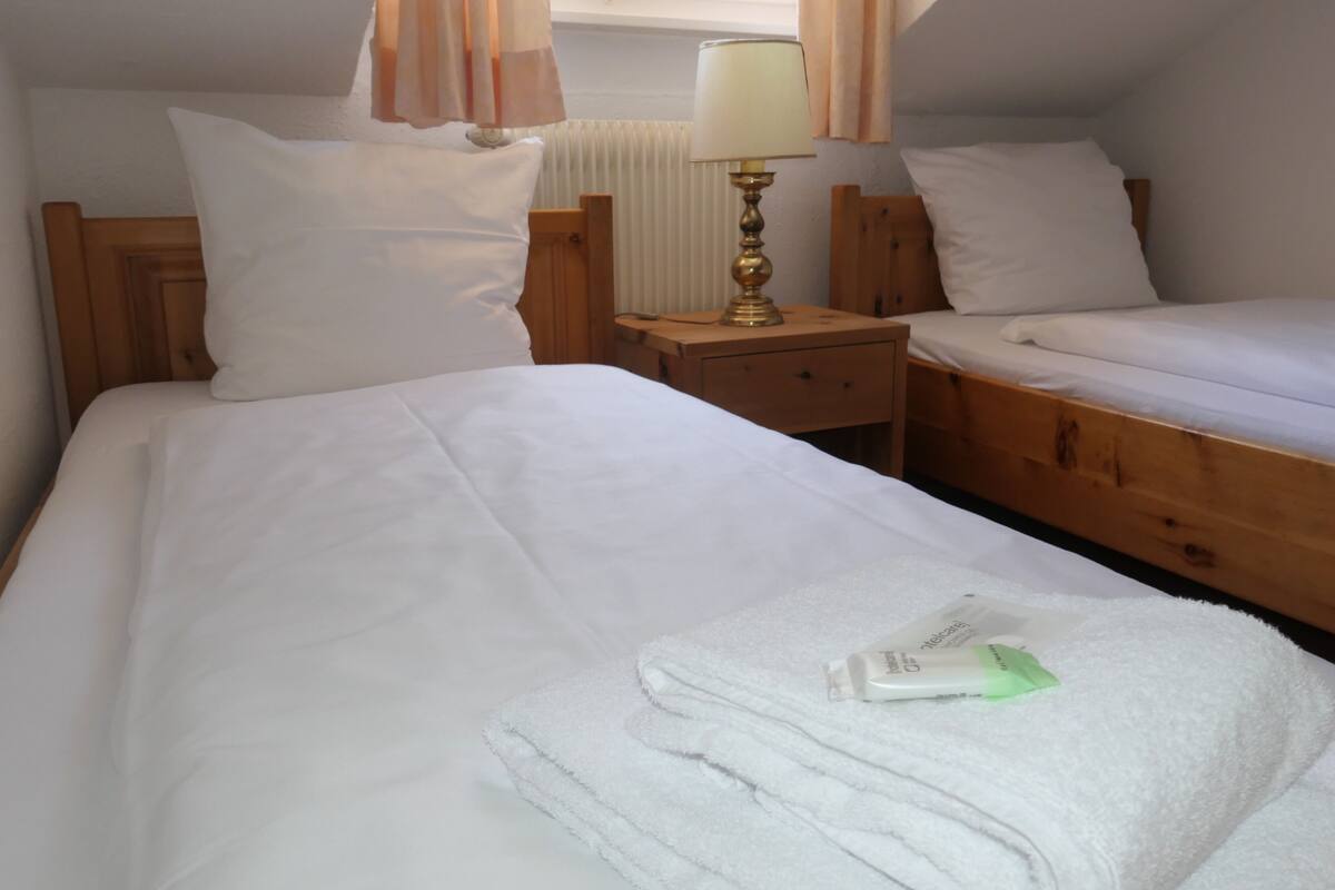Quarto de hotel com duas camas, uma mesinha de cabeceira com abajur e uma janela com cortinas.