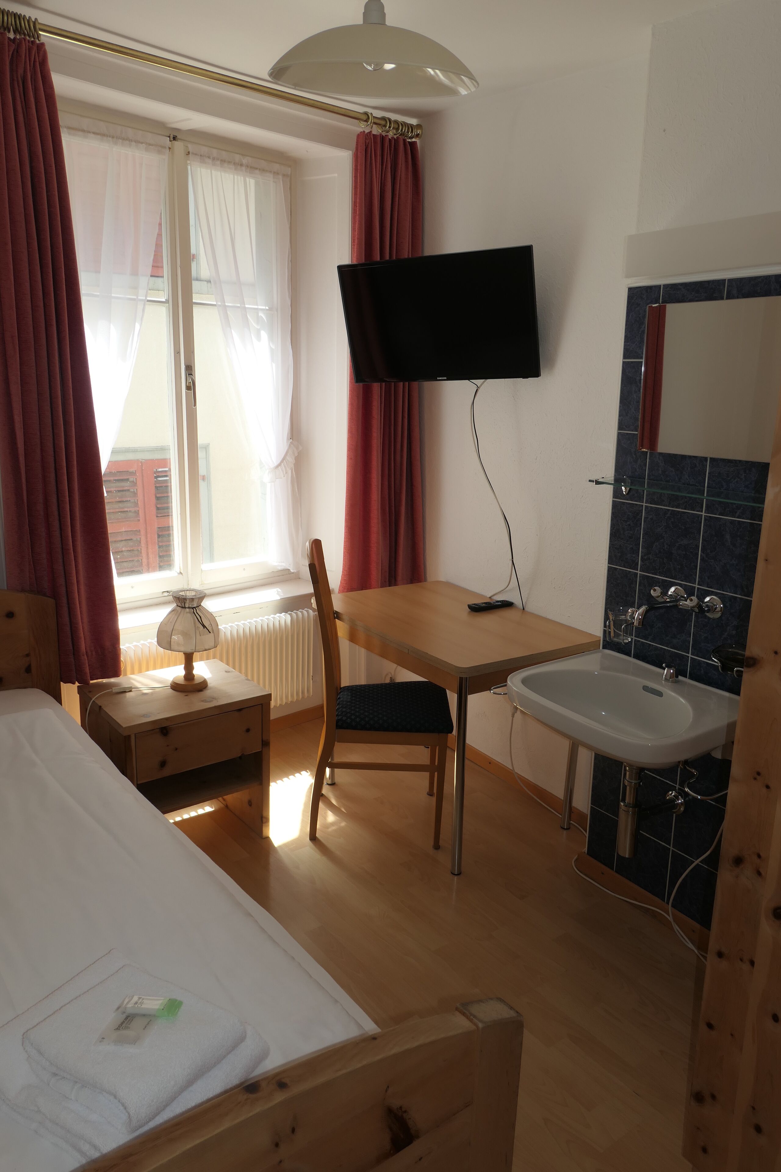 A hotel room with a bed, a desk with a chair, a television, a window with curtains, a radiator, and a sink.