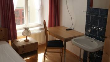 Habitación de hotel con cama, escritorio con silla, televisión, ventana con cortinas, radiador y lavamanos.