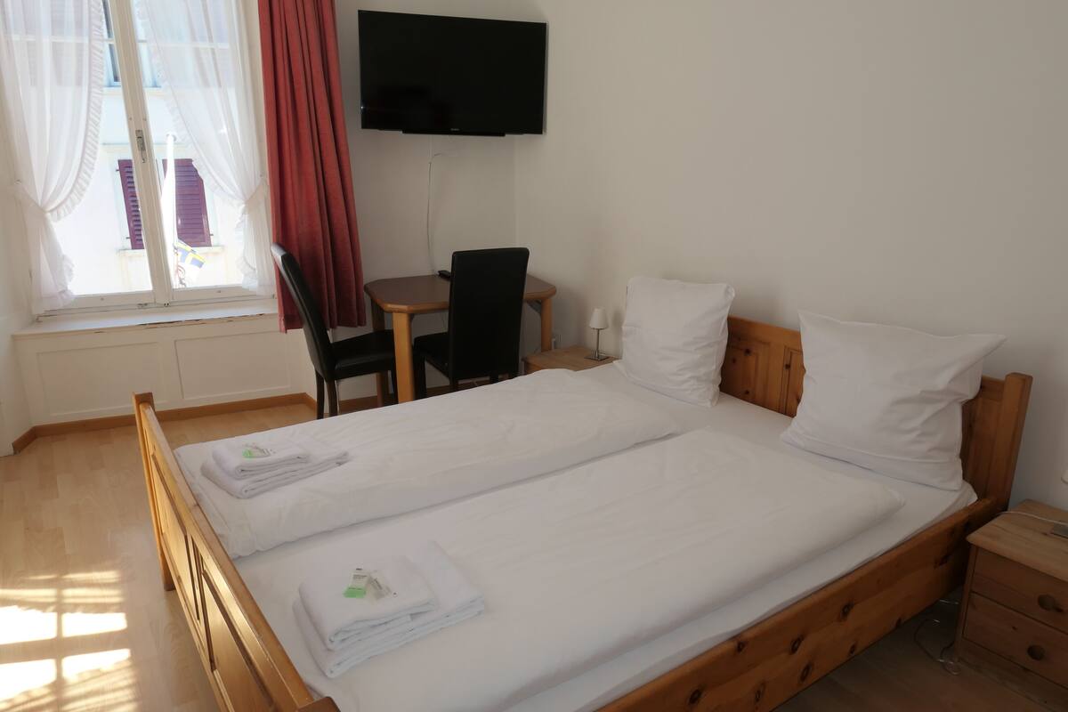 Quarto de hotel com cama de madeira, escrivaninha com cadeira, televisão na parede e janela com cortinas.