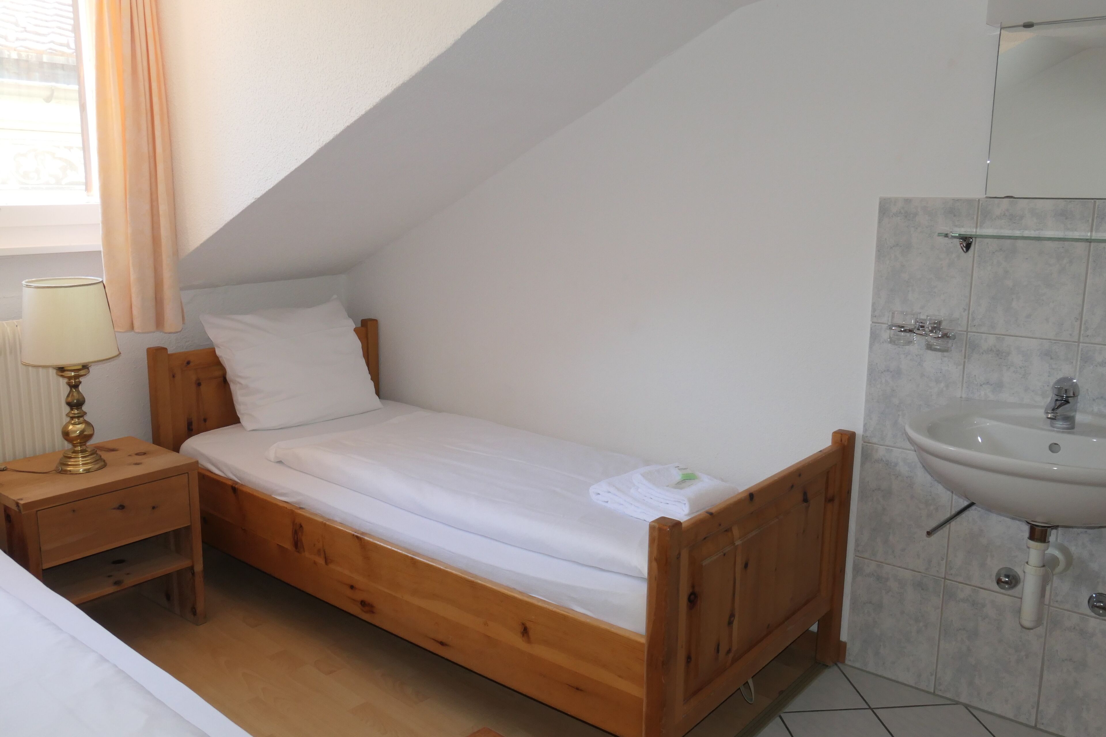 A single bed with a wooden frame, a bedside table with a lamp, and a bathroom with a sink and mirror.