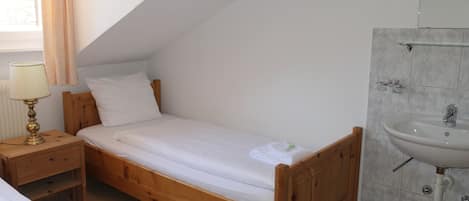 A single bed with a wooden frame, a bedside table with a lamp, and a bathroom with a sink and mirror.