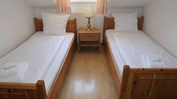 A room with two wooden beds, each with white bedding and pillows, a small wooden nightstand with a lamp between them, and a window with curtains.