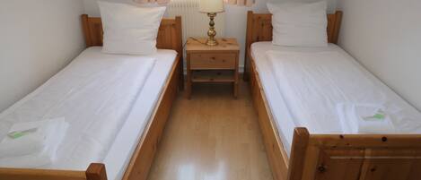 A room with two wooden beds, each with white bedding and pillows, a small wooden nightstand with a lamp between them, and a window with curtains.