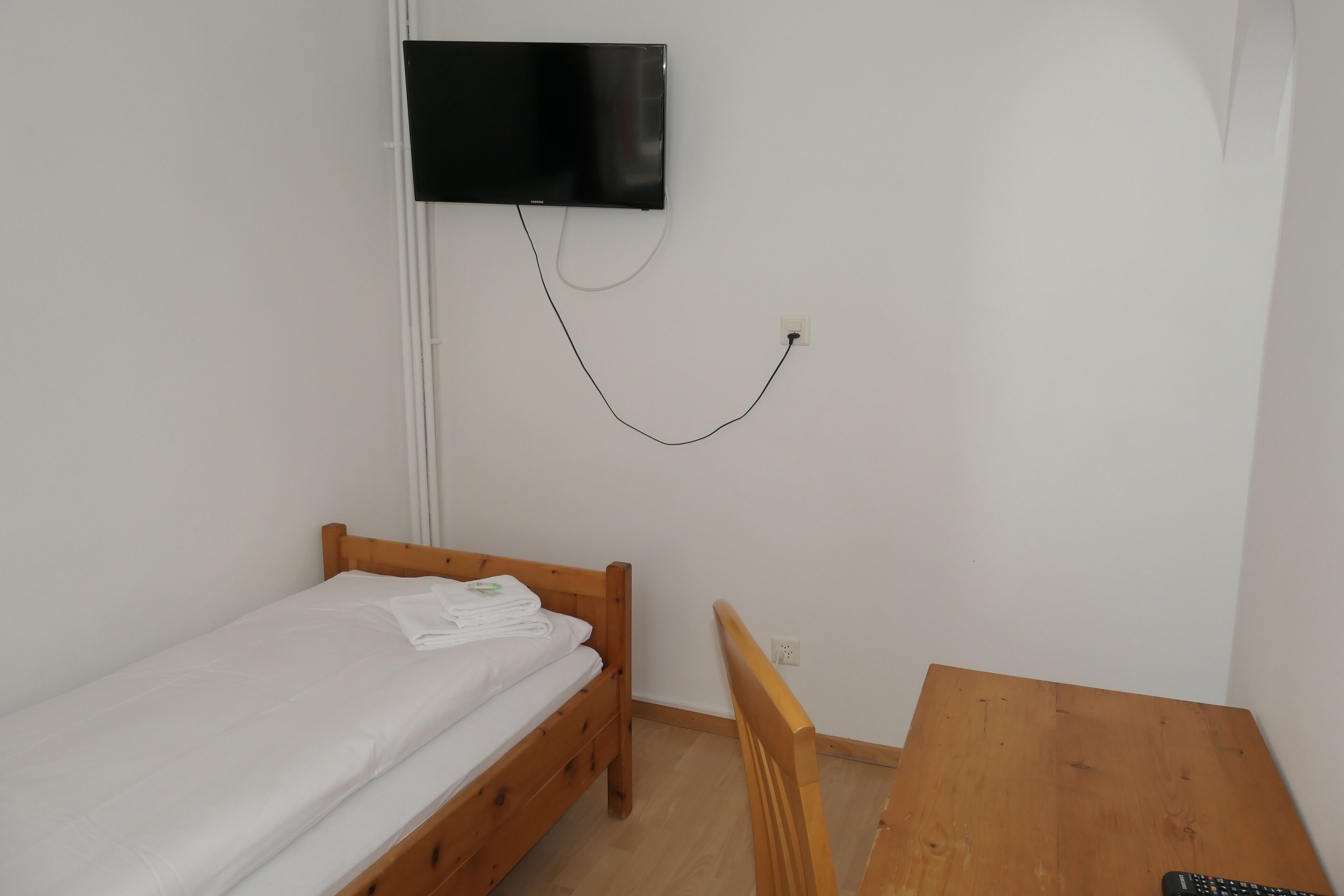 A small, simple room with a single bed, a wooden table, and a mounted TV.