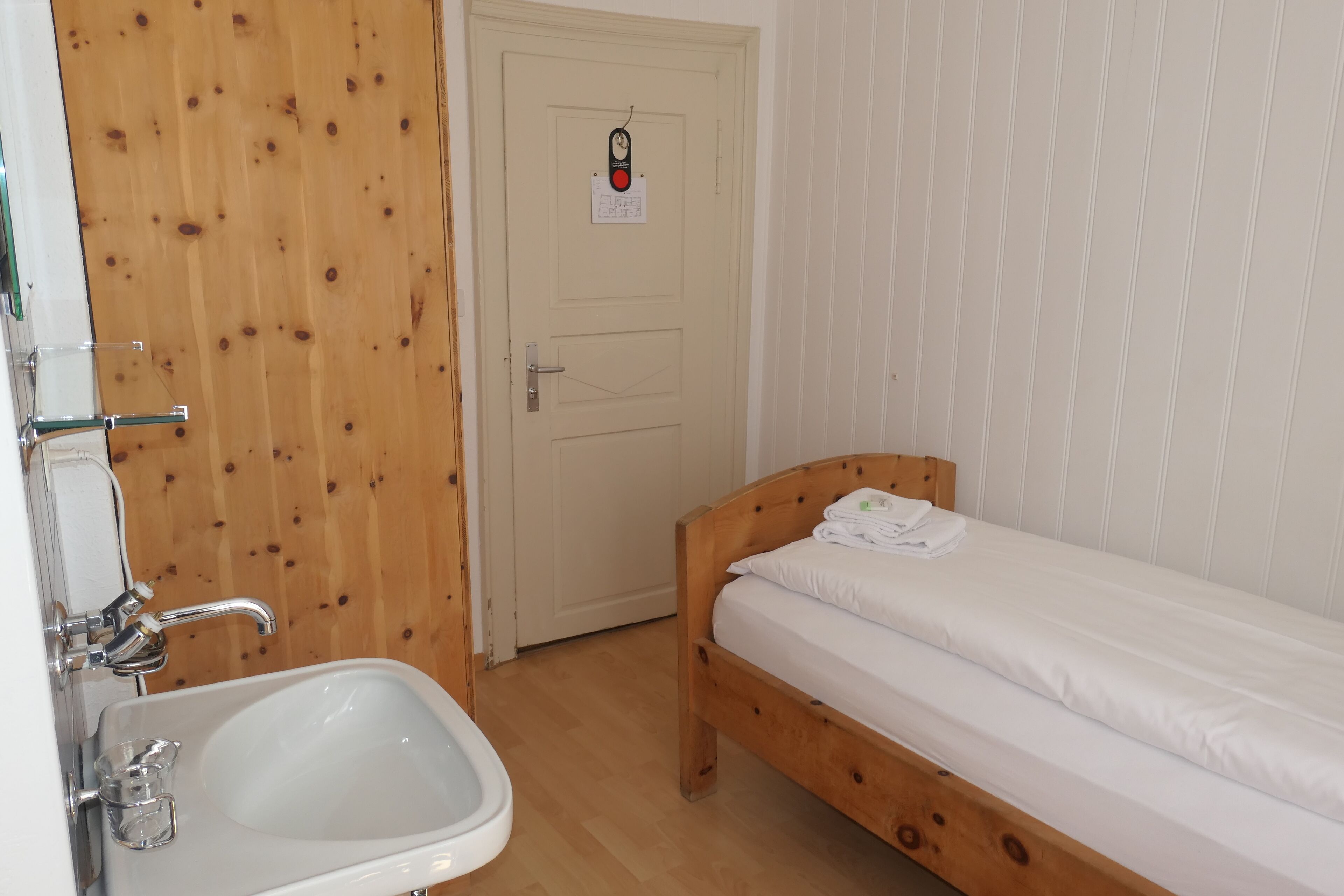 Una stanza piccola e pulita con una porta in legno, un lavandino bianco e un letto singolo con biancheria da letto bianca.