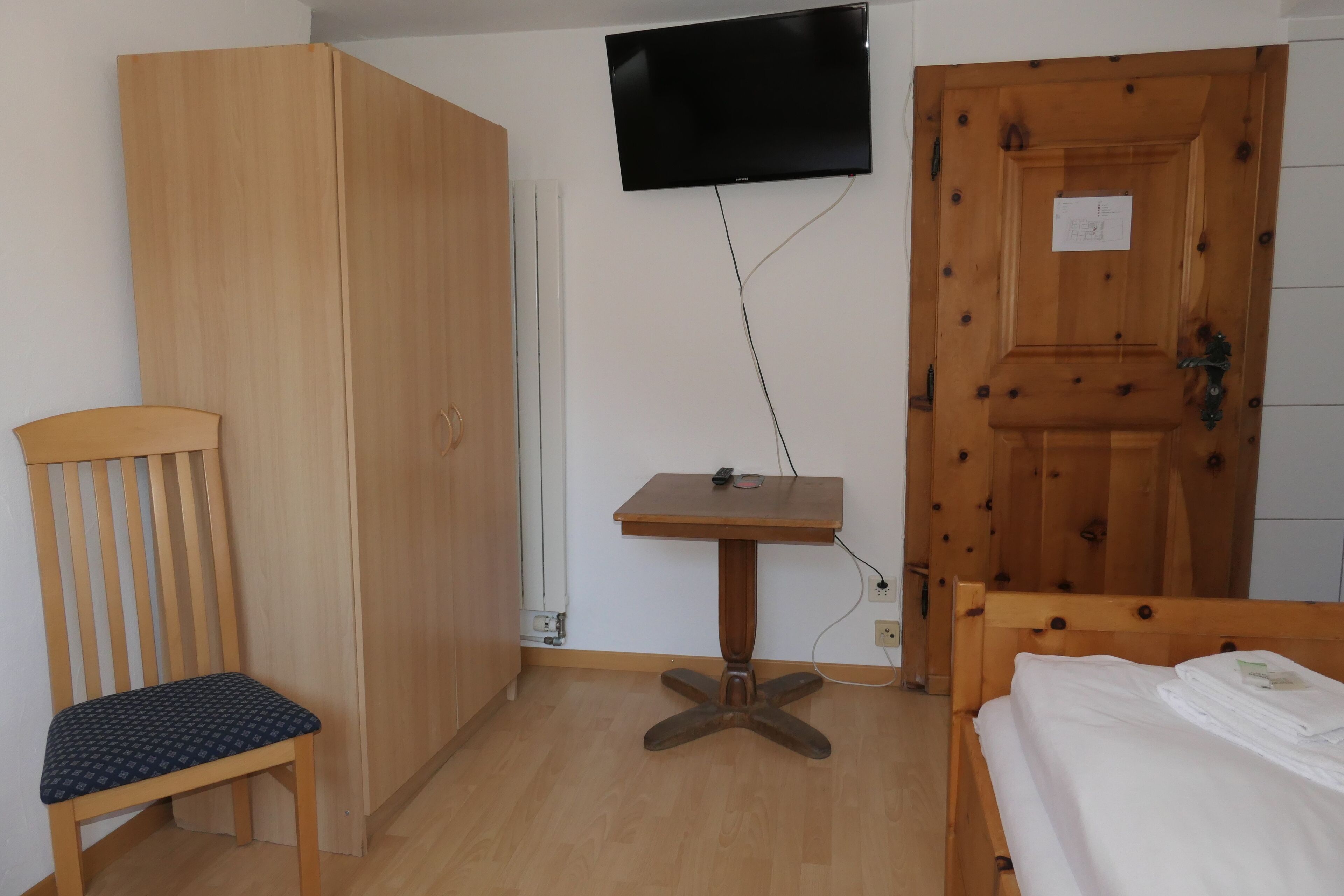 Een kamer met een houten garderobe, een aan de muur bevestigde televisie, een klein houten tafeltje, een stoel en een bed met netjes opgemaakte witte lakens.