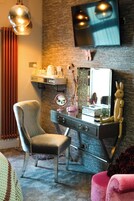 Quarto com toucador, cadeira, espelho, televisão e prateleira na parede.